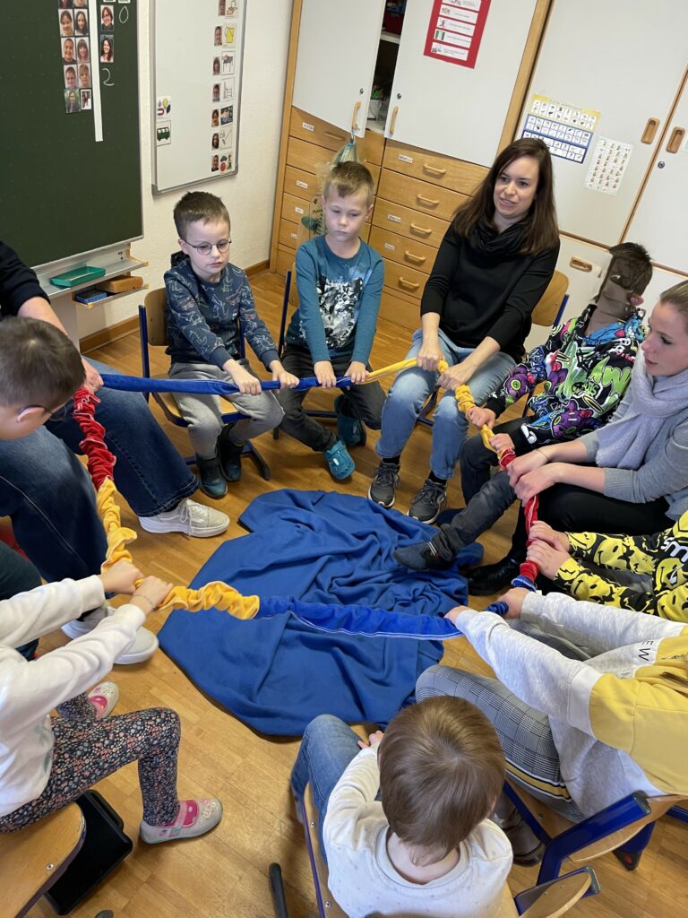 SchülerInen sitzen mit Lehrerinnen im Kreis und halten ein Band