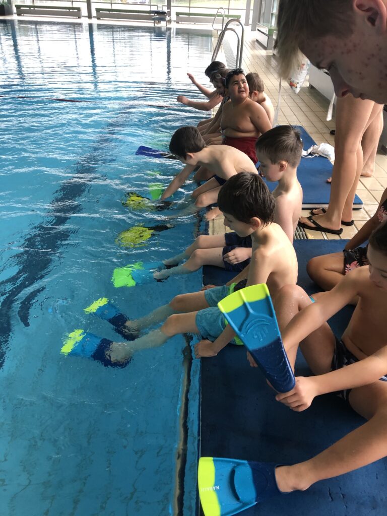 Schüler sitzen am Beckenrand des Schwimmbeckens und zeigen ihre Flossen an den Füßen 