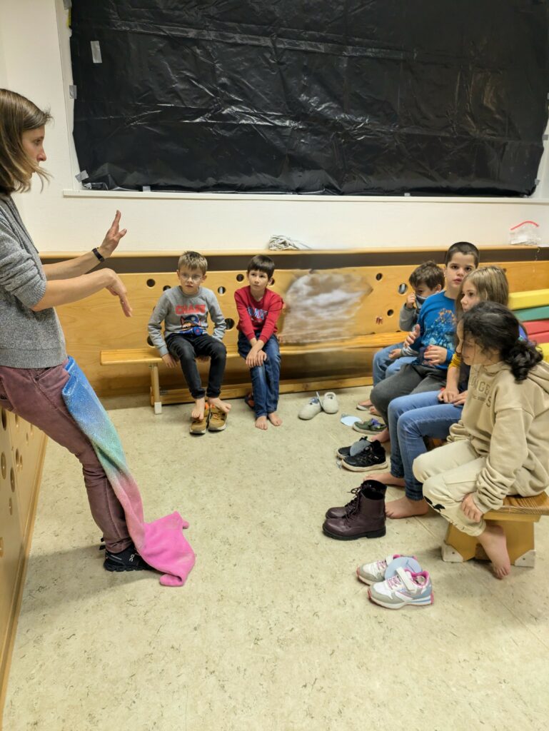 SchülerInnen sitzen auf Turnbänken und hören Lehrerin bei Erklärung über Meerjungfrauflossen zu